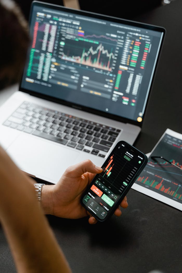 gallery-4 Person analyzing stock market data on a smartphone with a laptop displaying financial graphs in the background.